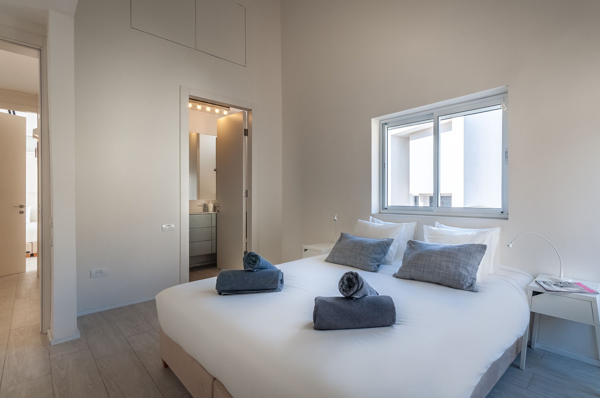 A modern hotel room with a large bed, two pillows, a bedside table with a lamp, and a window providing natural light.