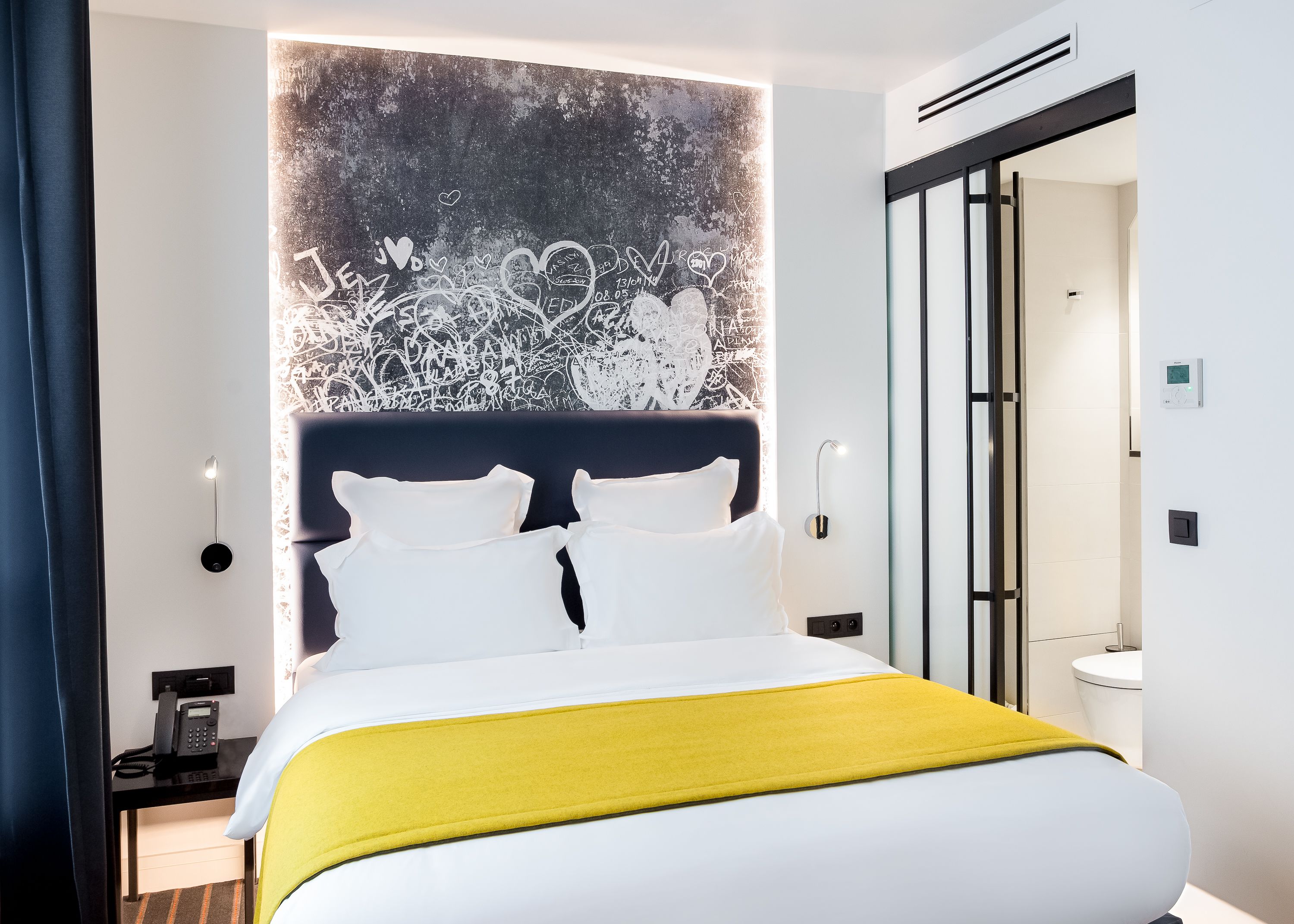 A modern hotel room with a large bed, a bedside table with a phone, and a wall-mounted light.