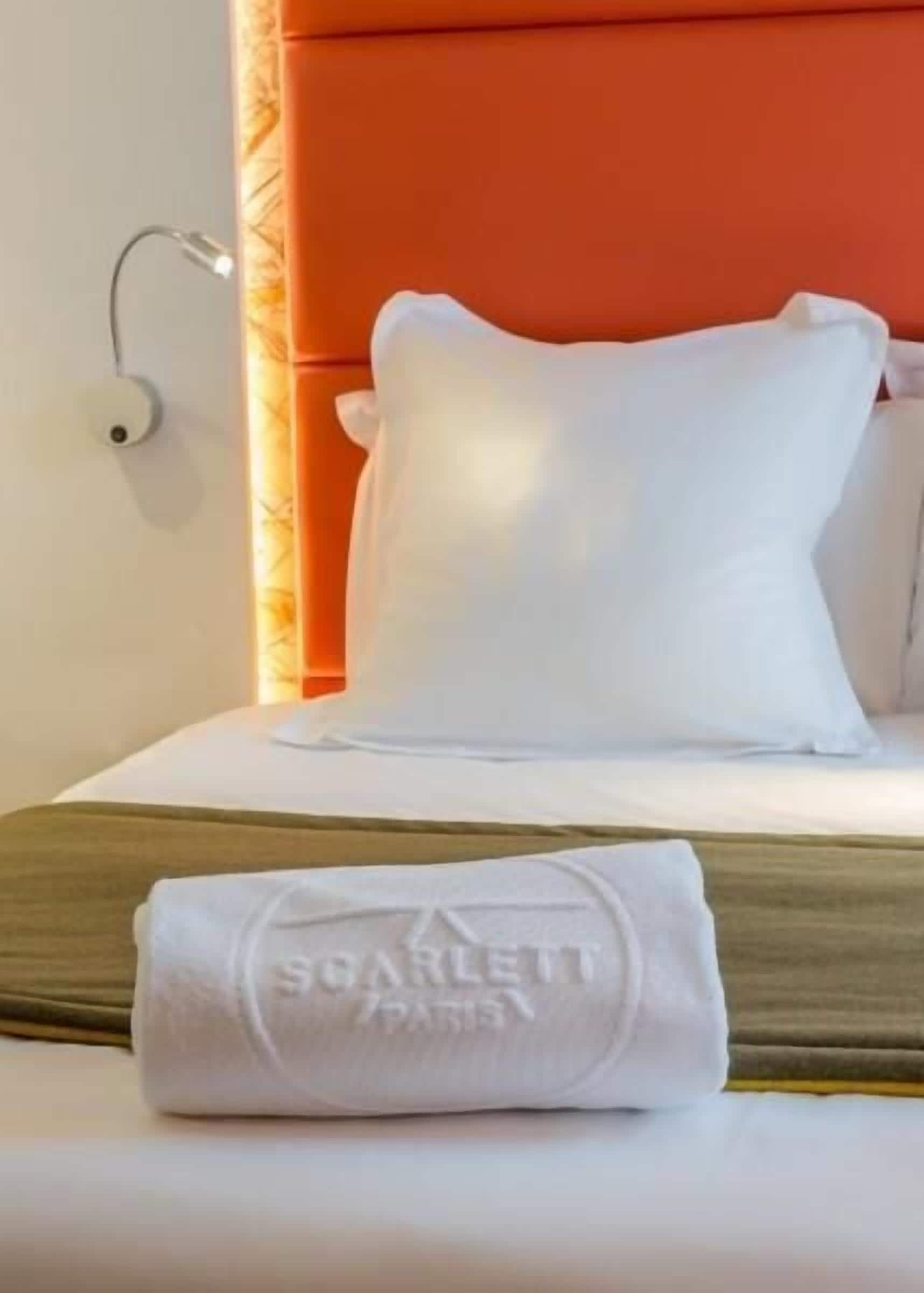 A neatly made bed with white bedding and a folded towel with "SOARLETT PARIS" text, a red headboard, and a wall-mounted light fixture.