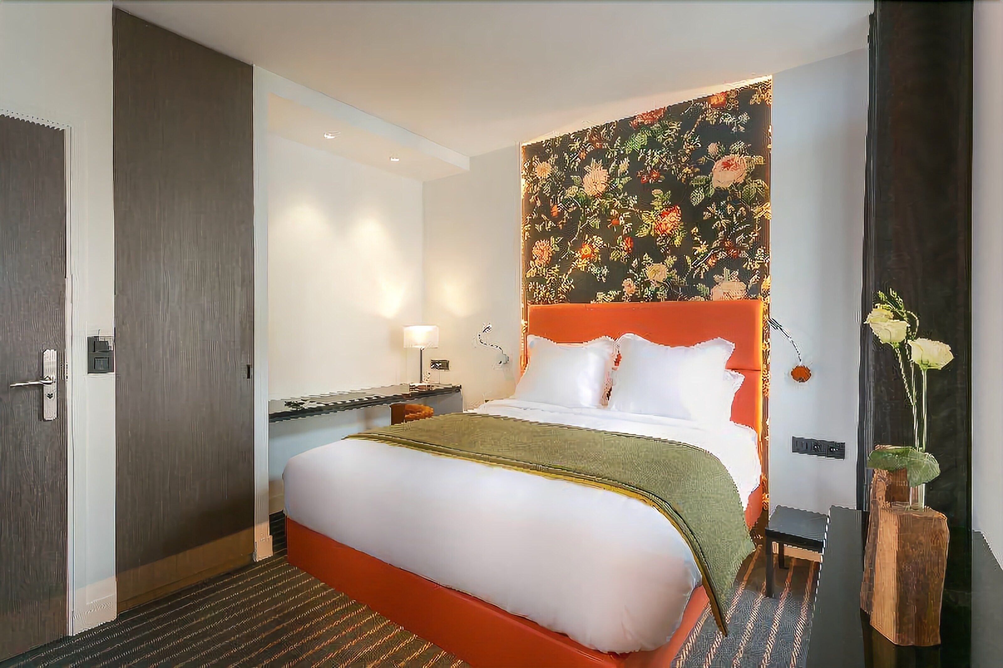 Una camera d'albergo con un letto grande, un comodino, una lampada e una testiera con motivo floreale.