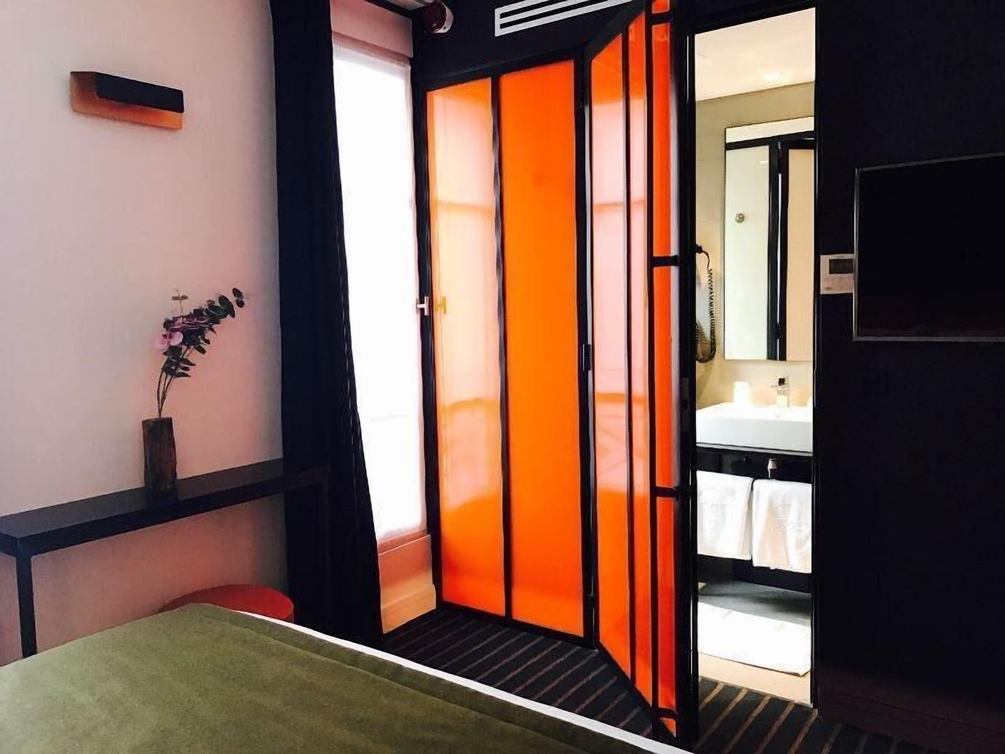 A hotel room with a bed, a small table, a vase with flowers, and a sliding door leading to a bathroom.