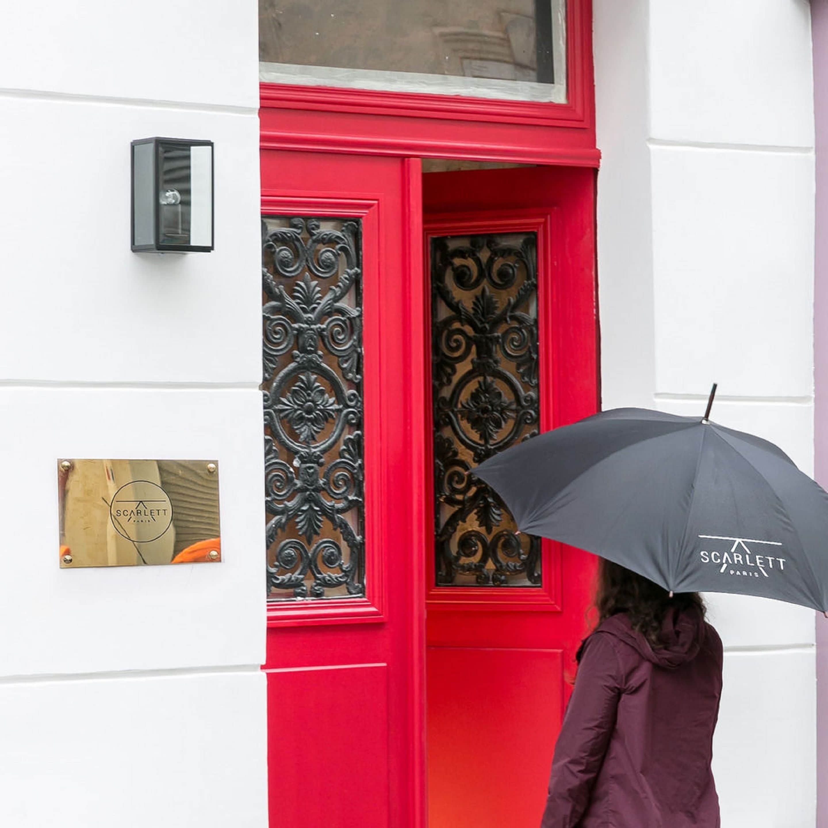 Uma pessoa com guarda-chuva preto em frente a uma porta vermelha com portão de metal decorativo.
