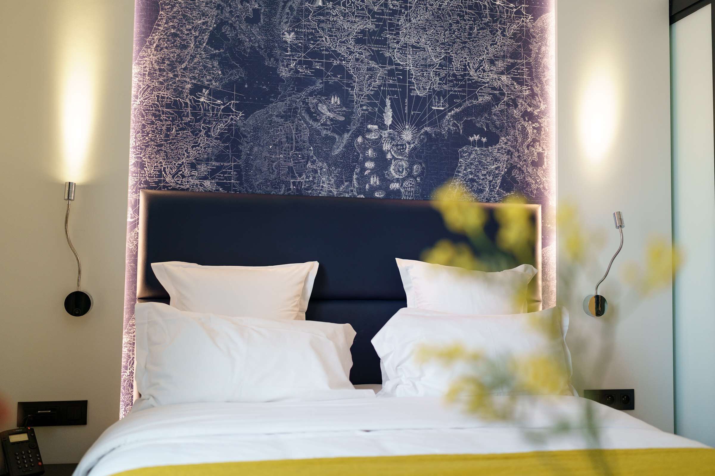 Un lit bien fait, avec du linge de lit blanc et un élément décoratif jaune, une tête de lit sombre et une grande carte du monde peinte sur le mur derrière.