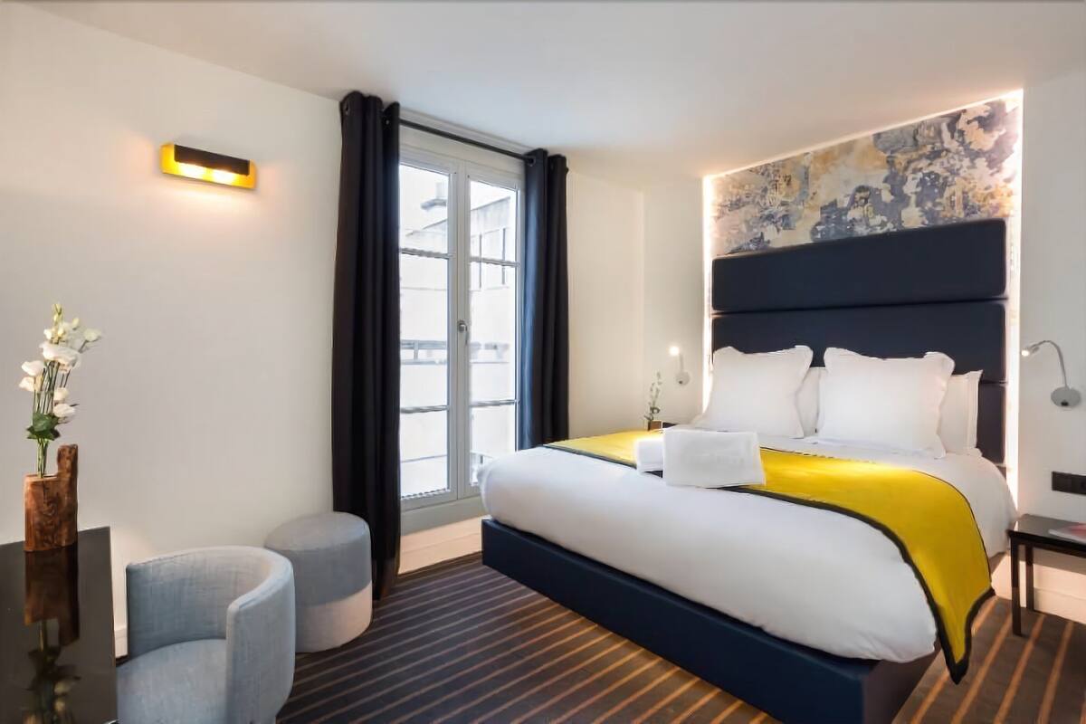 A hotel room with a large bed, a small round table, a chair, a vase with flowers, and a window with curtains.