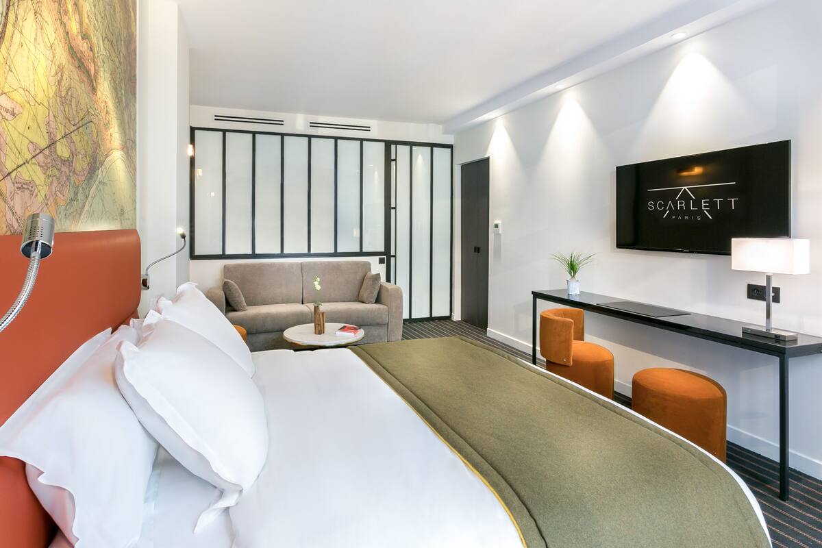 A hotel room with a bed, a sofa, a desk, and a TV displaying 'SCARLETT PARIS'.