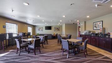 A hotel dining area with multiple tables and chairs, a kitchen area, and a television on the wall.