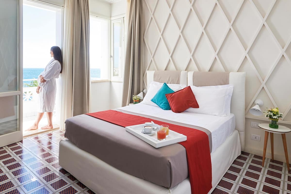 A hotel room with a bed, a tray with breakfast items, a window with a view of the sea, and a person standing by the window.