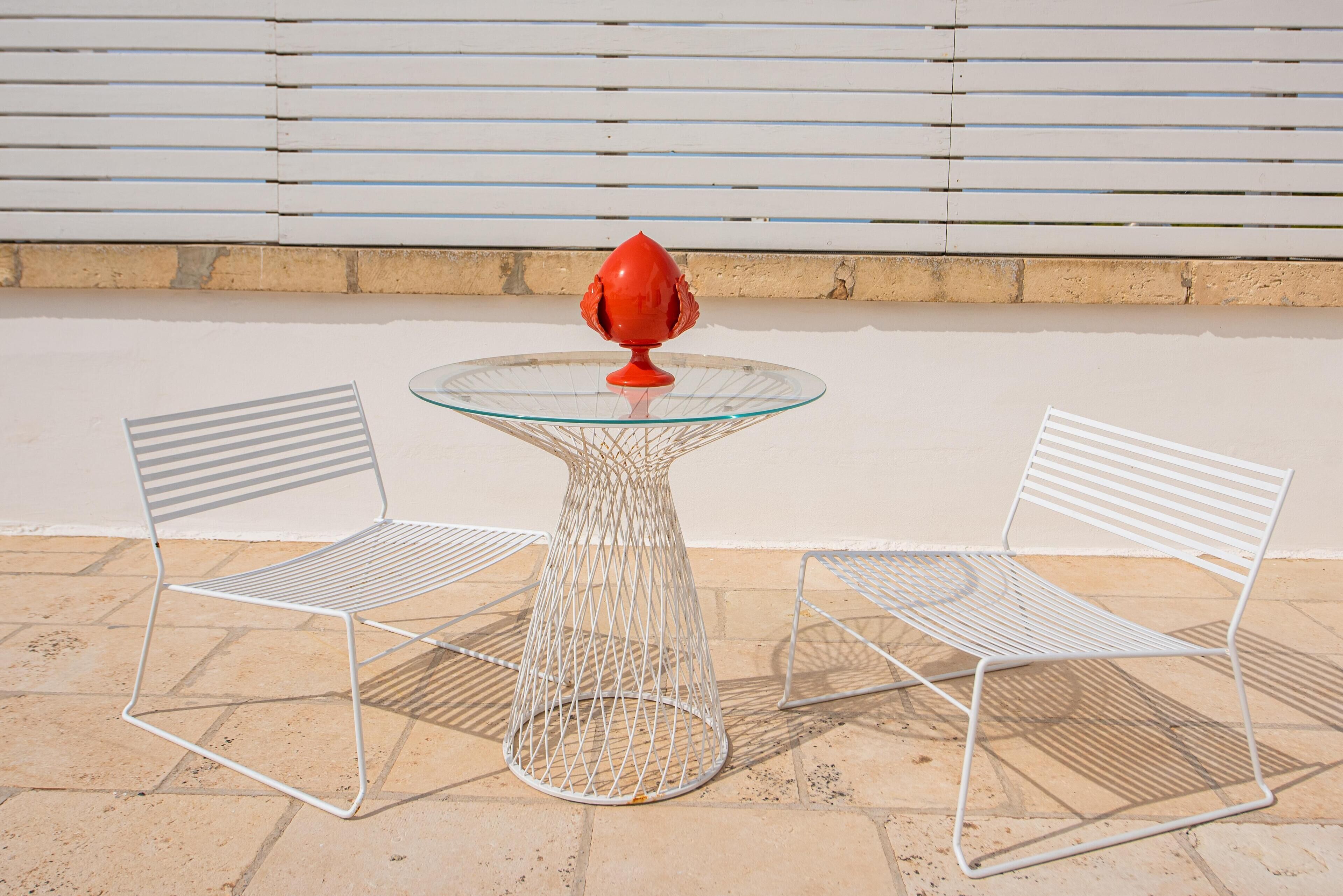 A table with a glass top and a red decorative item, flanked by two white metal chairs, set on a tiled patio.