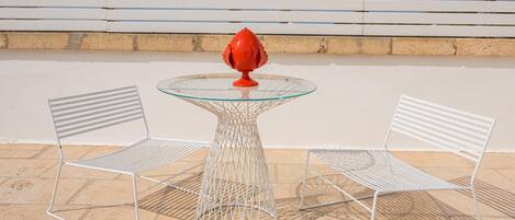 A table with a glass top and a red decorative item, flanked by two white metal chairs, set on a tiled patio.