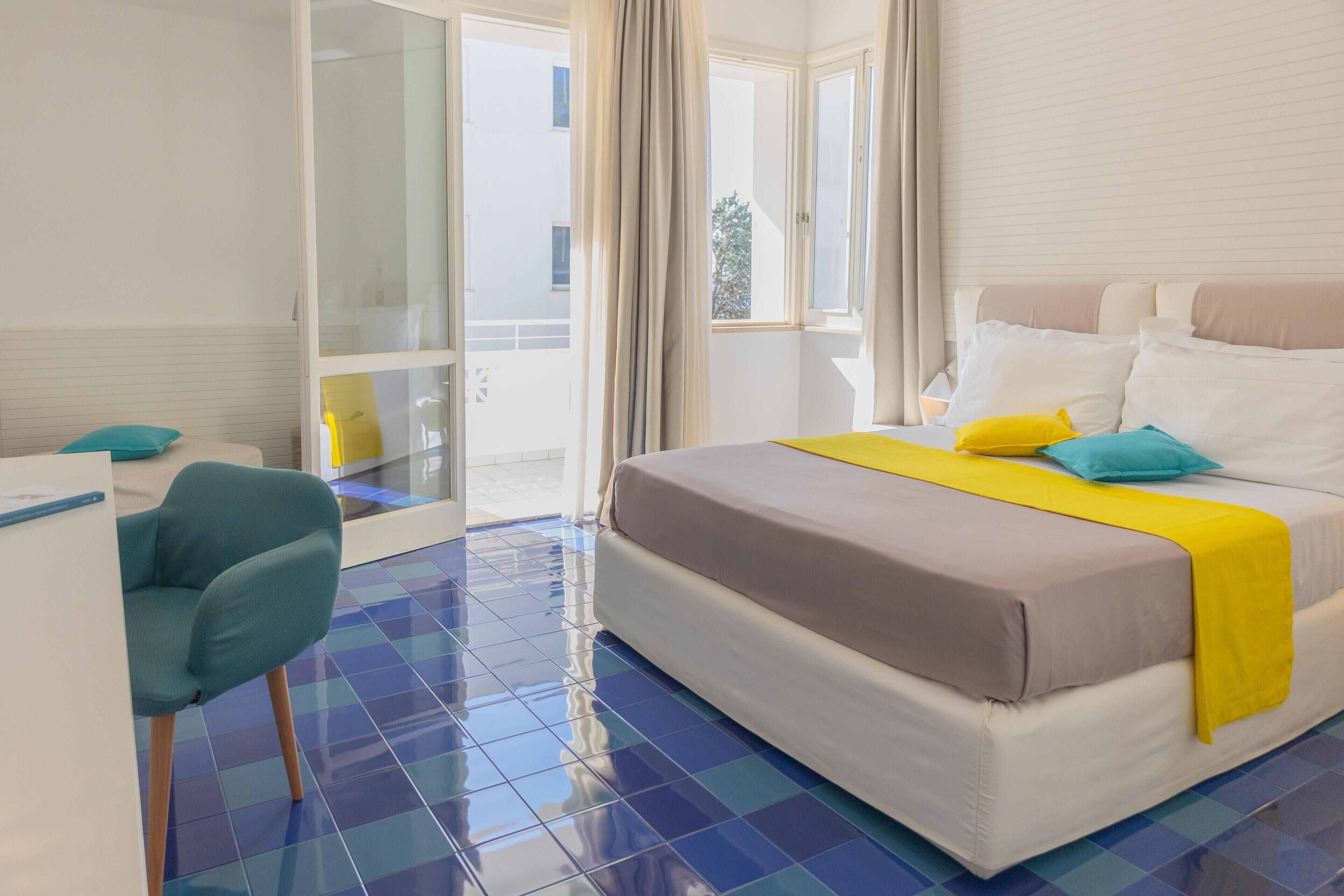 A hotel room with a bed, a desk, a chair, and a balcony with curtains.