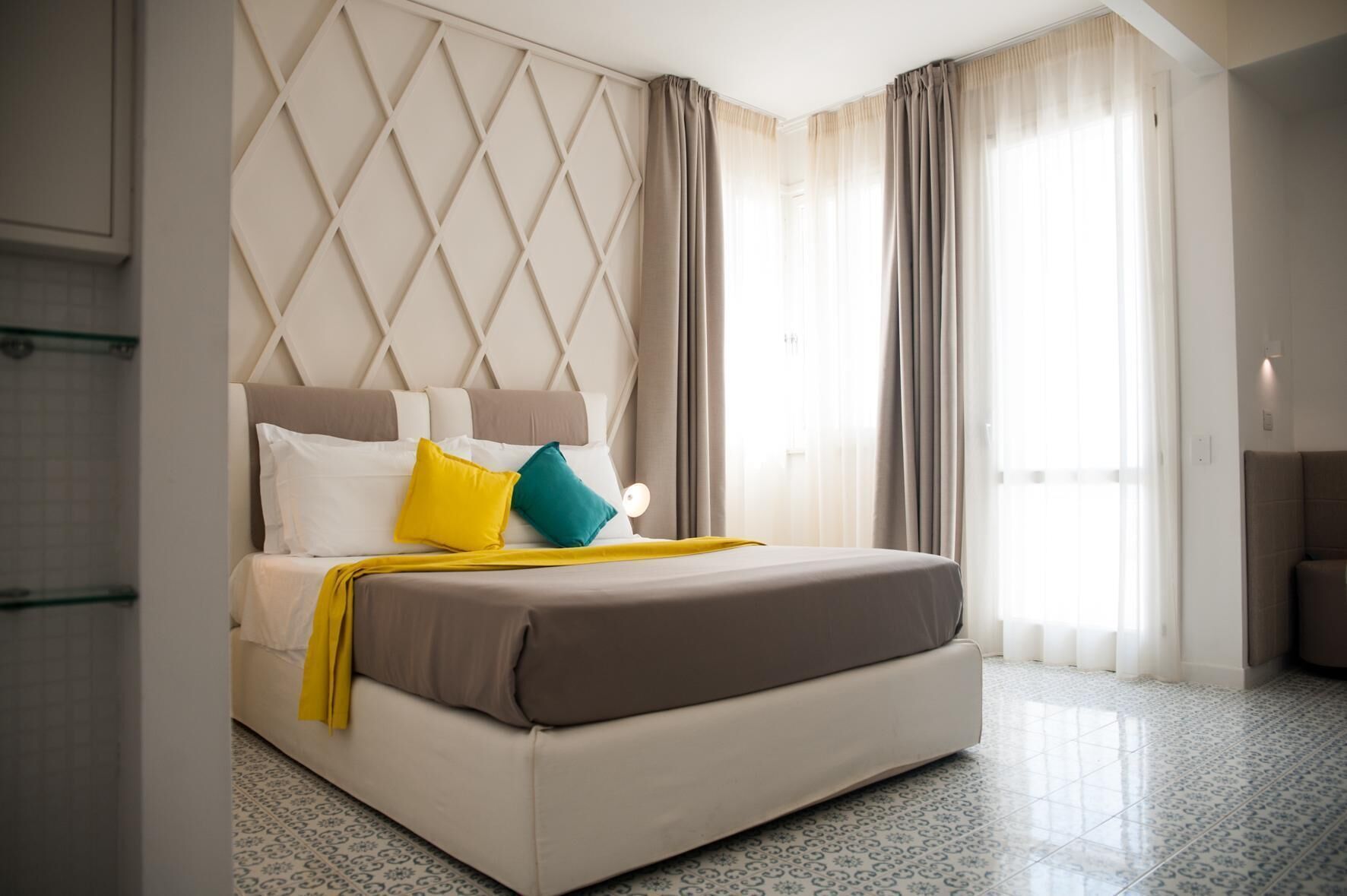 A modern hotel room with a large bed, a headboard with a geometric pattern, and a view of the outdoors through large windows.