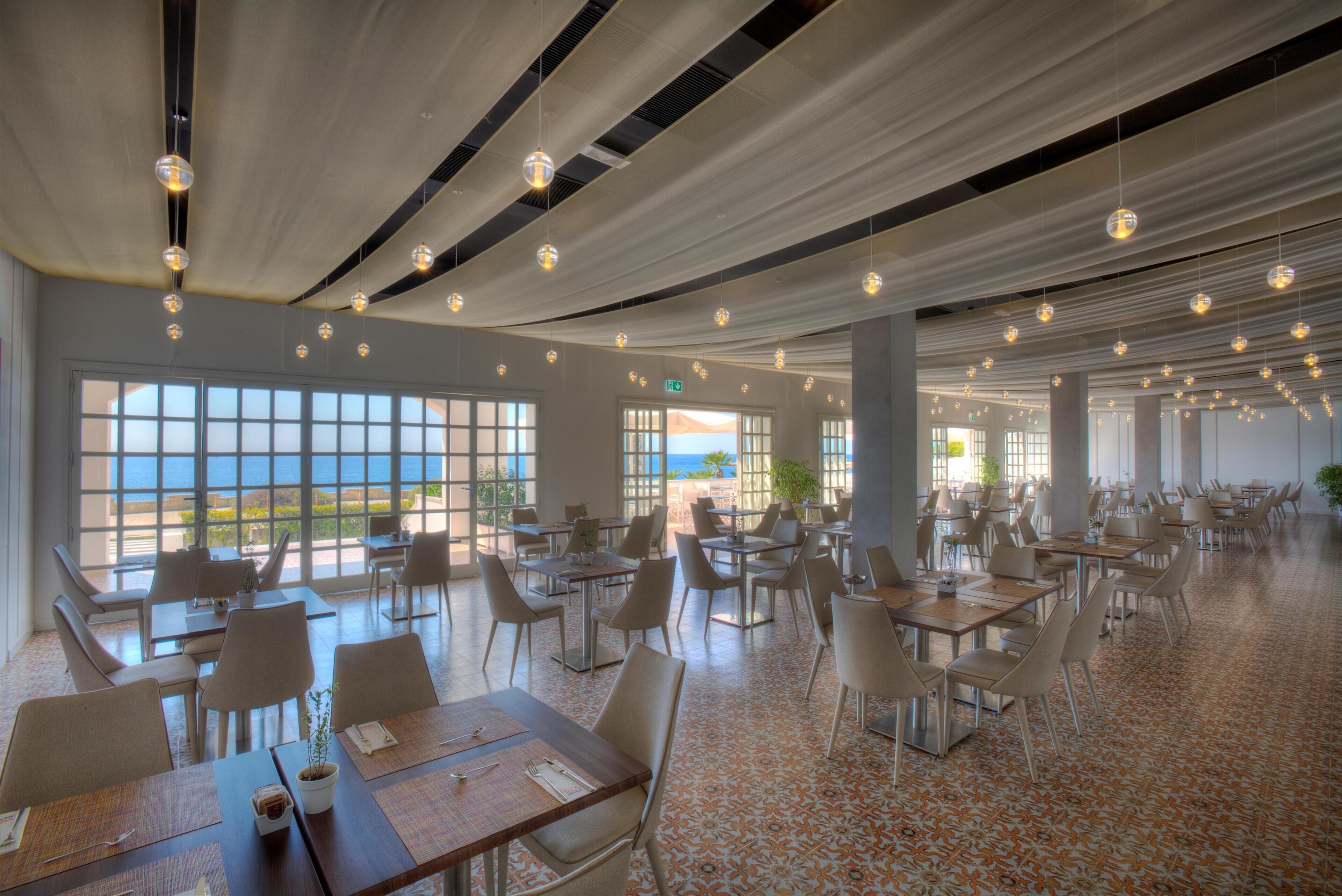 Un vaste espace dédié à la salle à manger, avec de nombreuses tables et chaises, de grandes fenêtres offrant une vue sur la mer et des luminaires sphériques suspendus.
