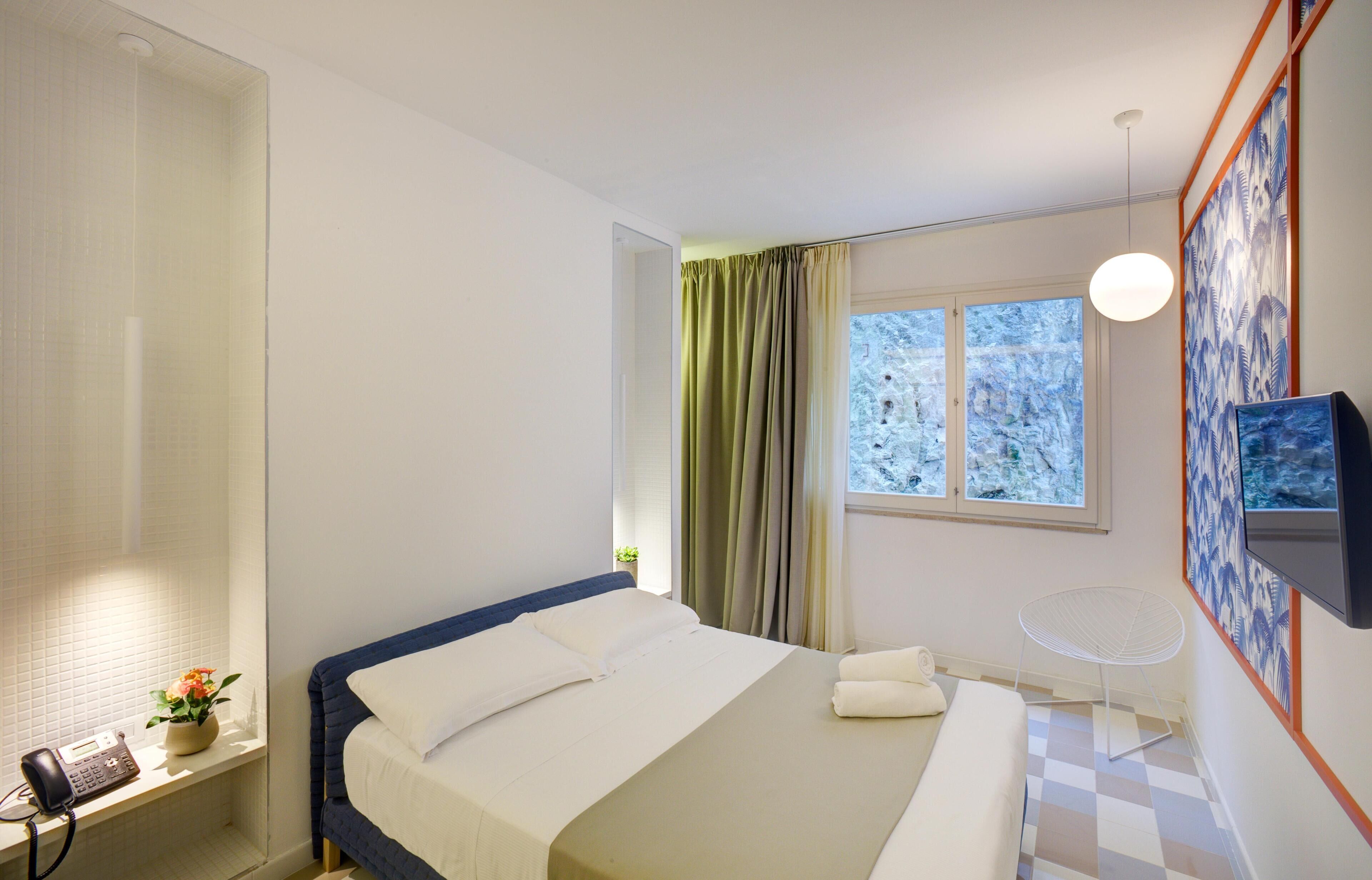 Quarto de hotel com uma cama, uma escrivaninha com telefone, uma cadeira, uma televisão e uma janela com cortinas.