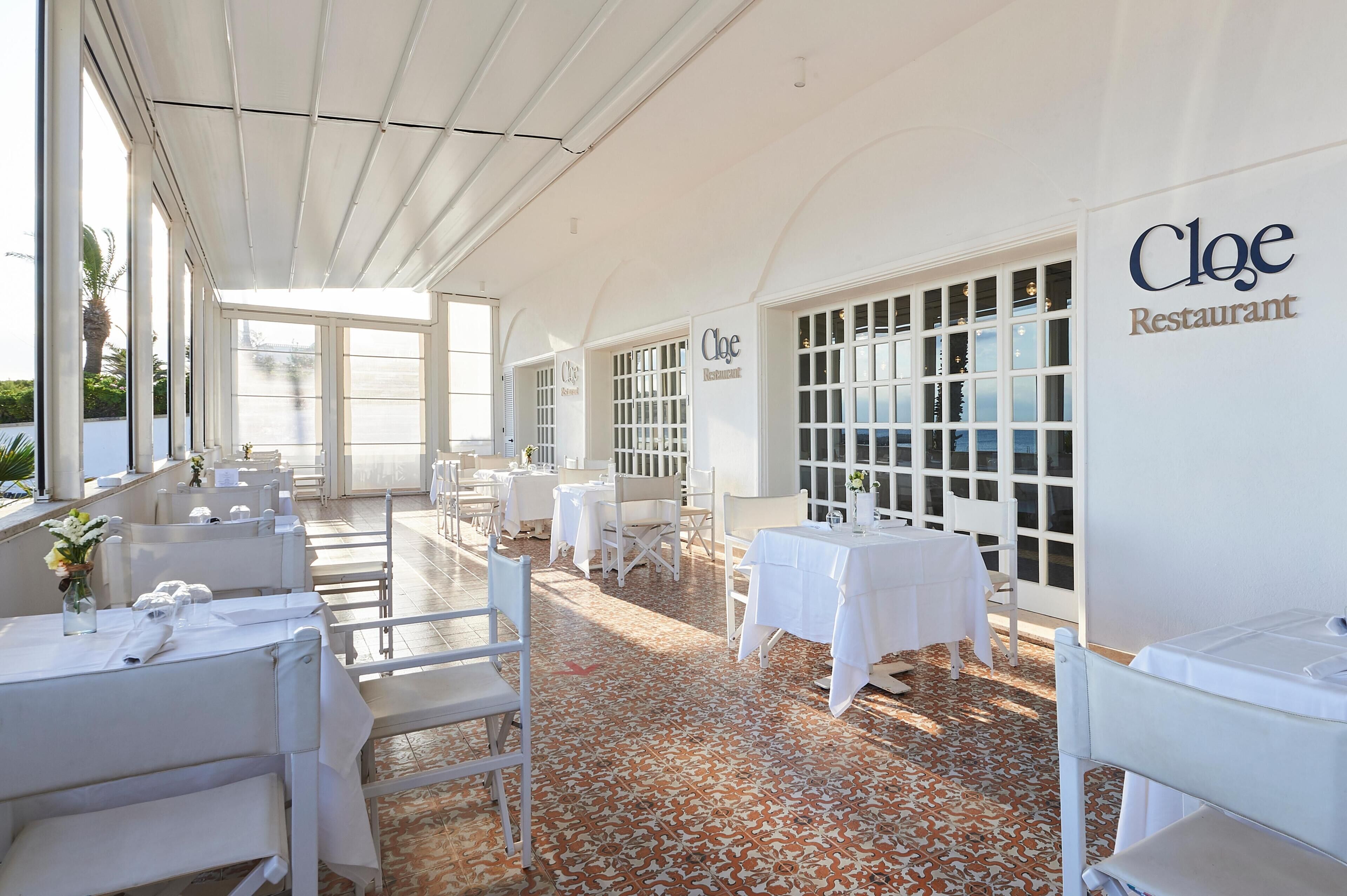 A restaurant with white tables and chairs, a tiled floor, and large windows.