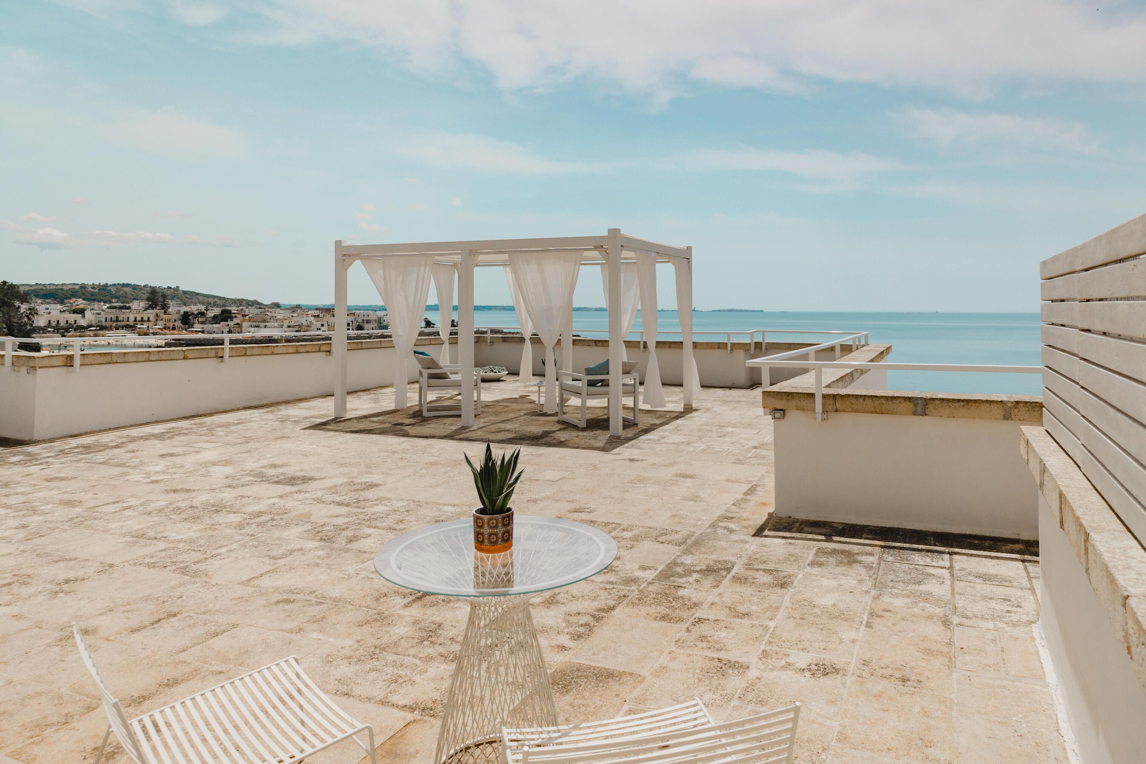 A rooftop terrace with a white pergola, a glass table with a potted plant, and white chairs.