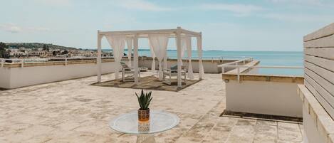 A rooftop terrace with a white pergola, a glass table with a potted plant, and white chairs.