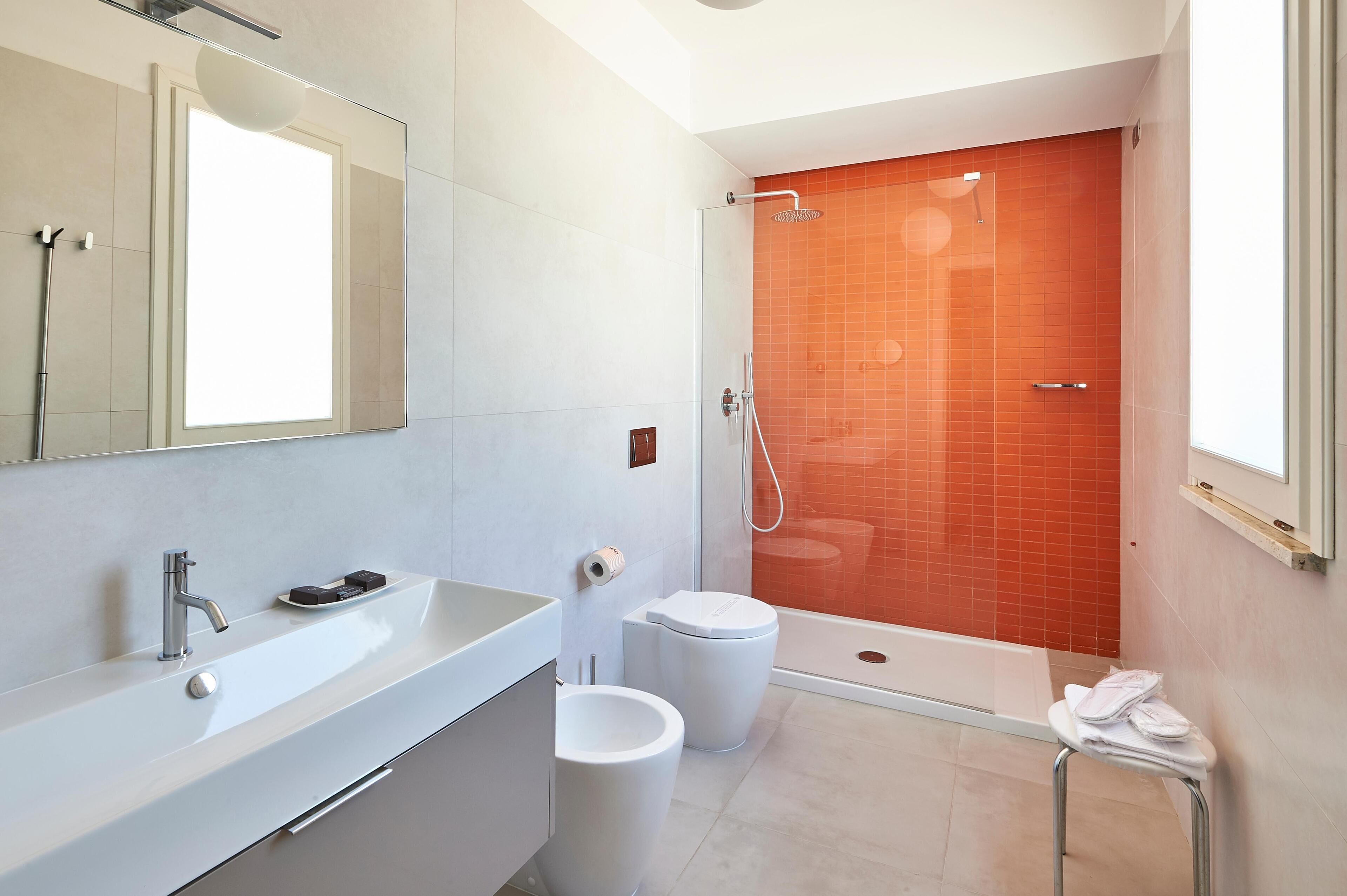 A modern bathroom with a large mirror, a white sink, two toilets, and a shower area with orange tiles.