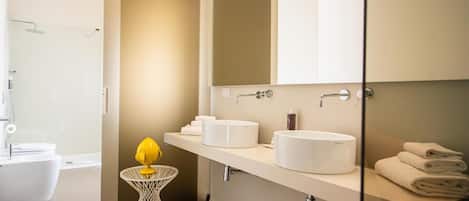 Un baño moderno con cabina de ducha de vidrio, un lavabo blanco con un espejo encima y una pequeña mesa redonda con un objeto amarillo.