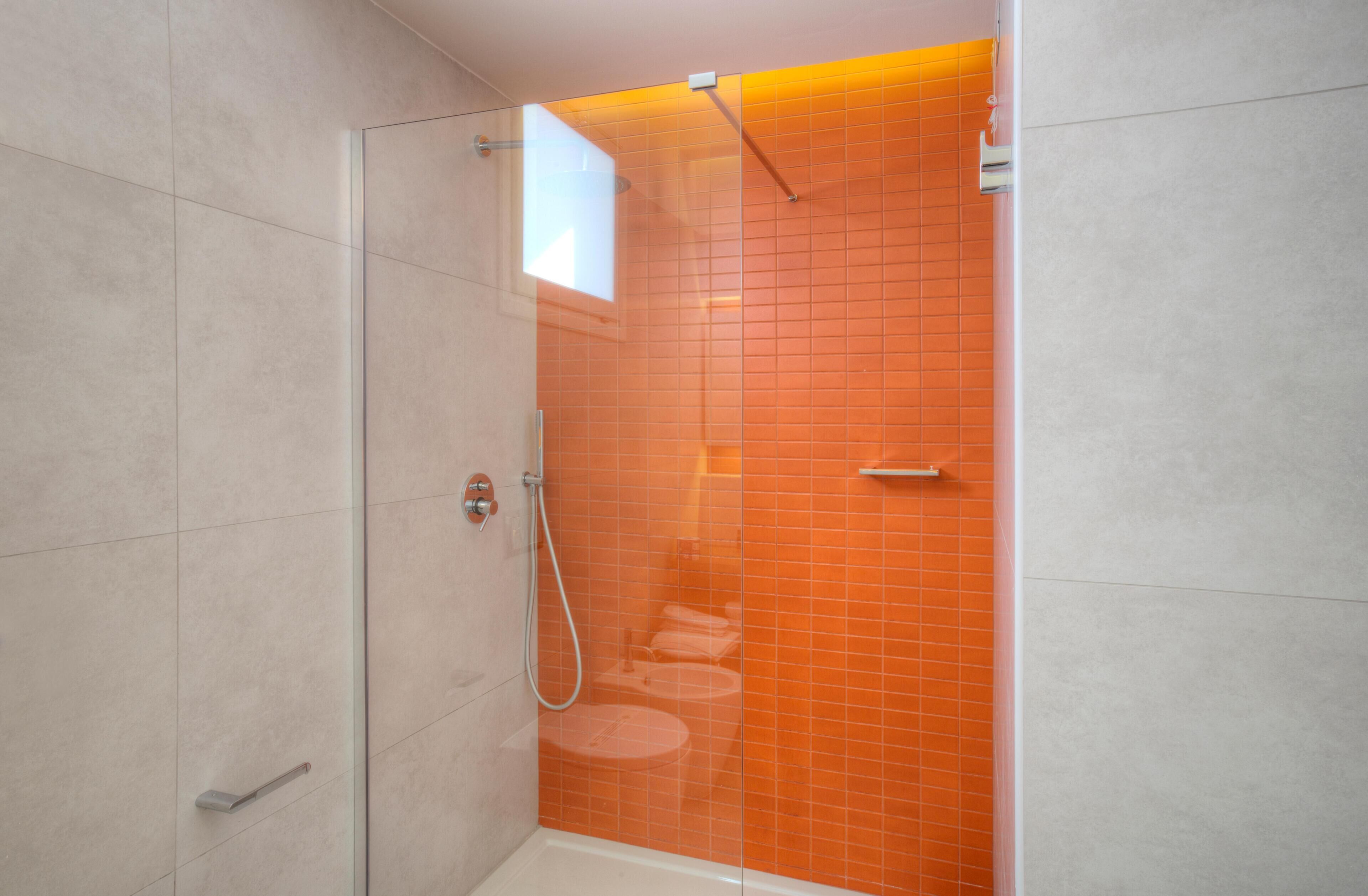 A modern bathroom with a glass-enclosed shower, white tiles, and an orange-tiled accent wall.
