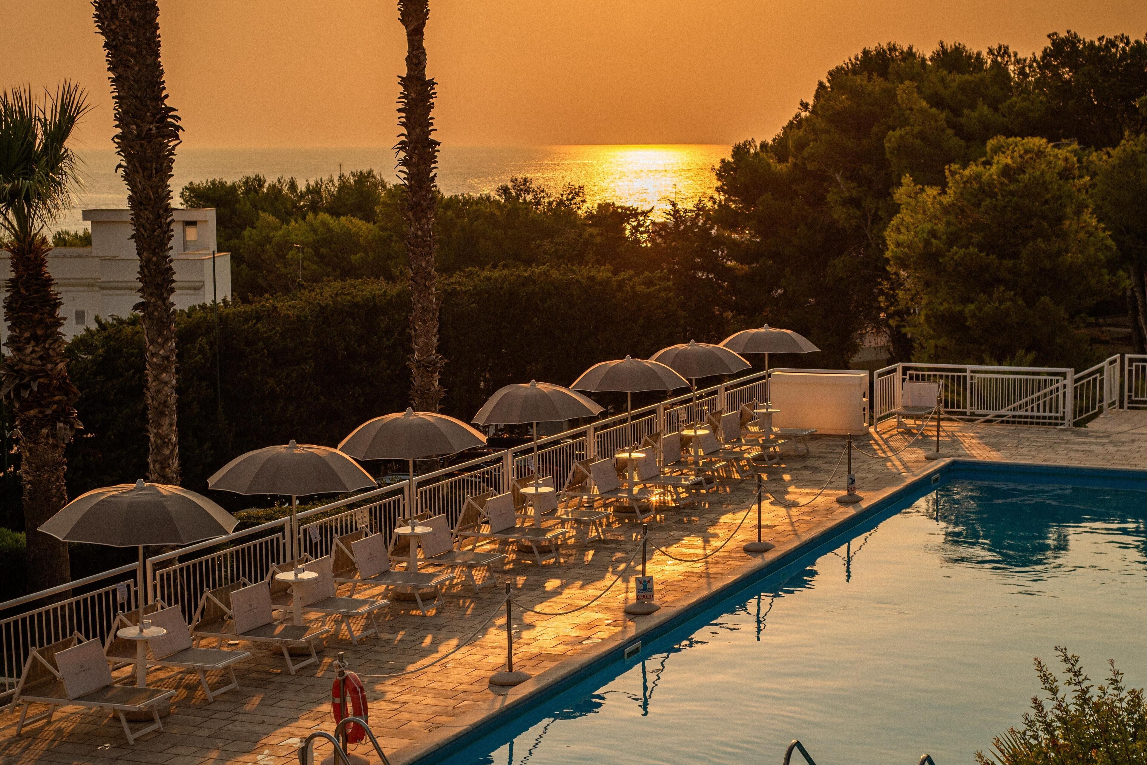 Zona de piscina con sillones de descanso y sombrillas, un flotador salvavidas y vista al atardecer sobre el mar.