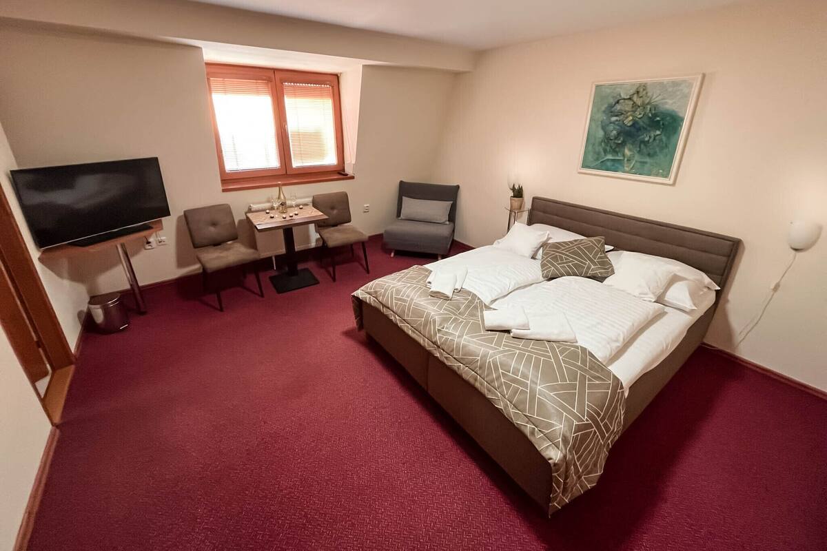 Quarto de hotel com cama, televisão, secretária e cadeiras, janela e um quadro na parede.