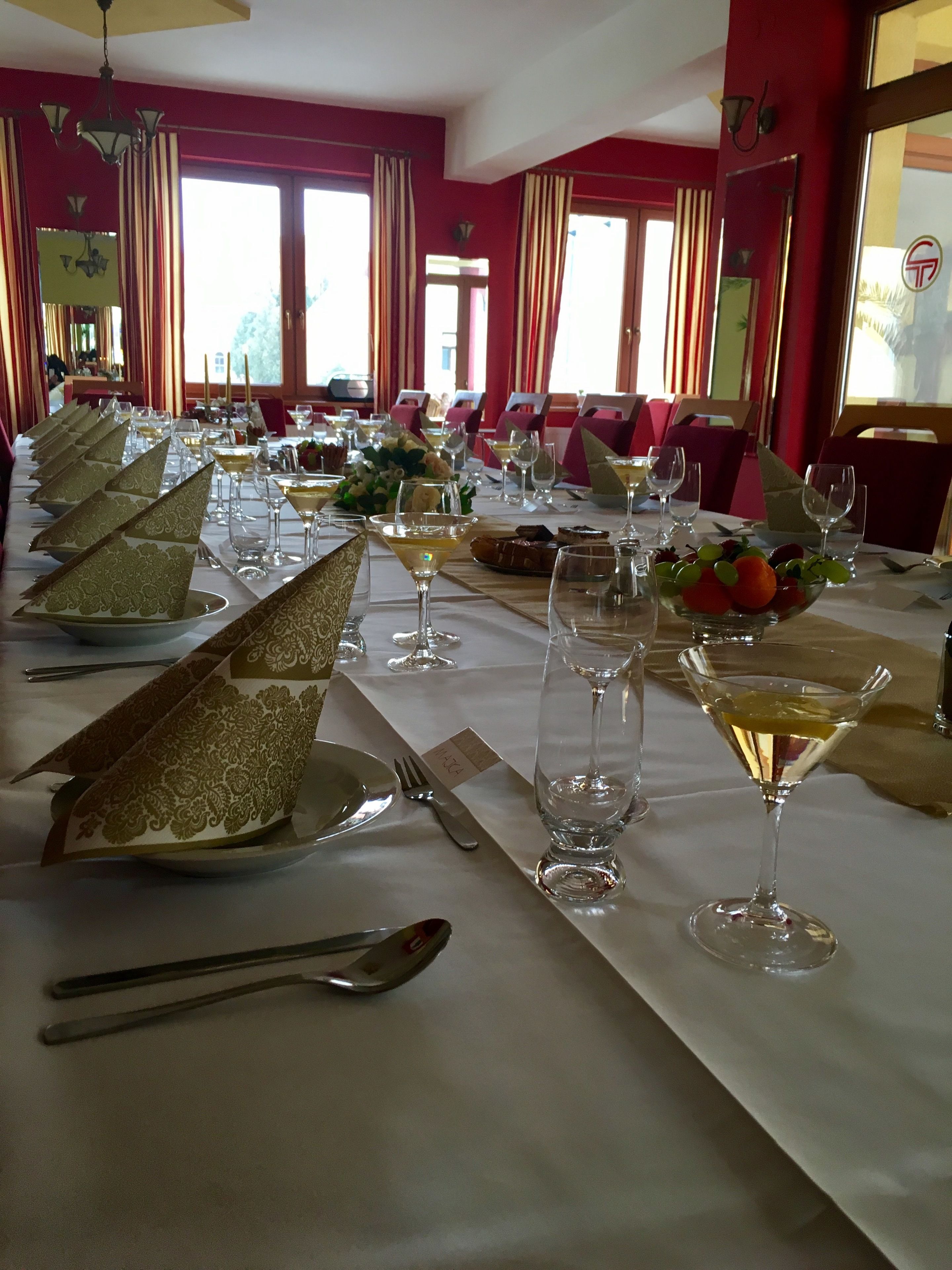 A formal dining room with a long table set for a meal, featuring gold-patterned napkins, wine glasses, and a fruit bowl.