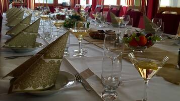 A formal dining room with a long table set for a meal, featuring gold-patterned napkins, wine glasses, and a fruit bowl.