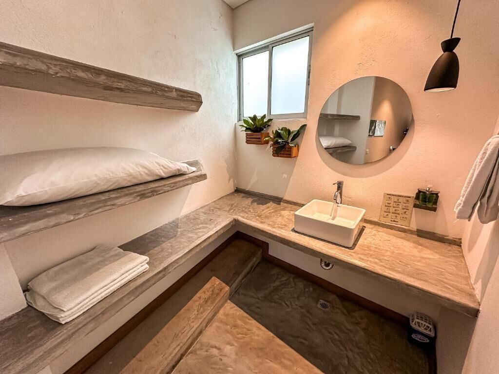 Ein kleines Badezimmer mit einem Holzwaschtisch, einem runden Spiegel, einem eingebauten Waschbecken und einem Fenster mit Pflanzen.