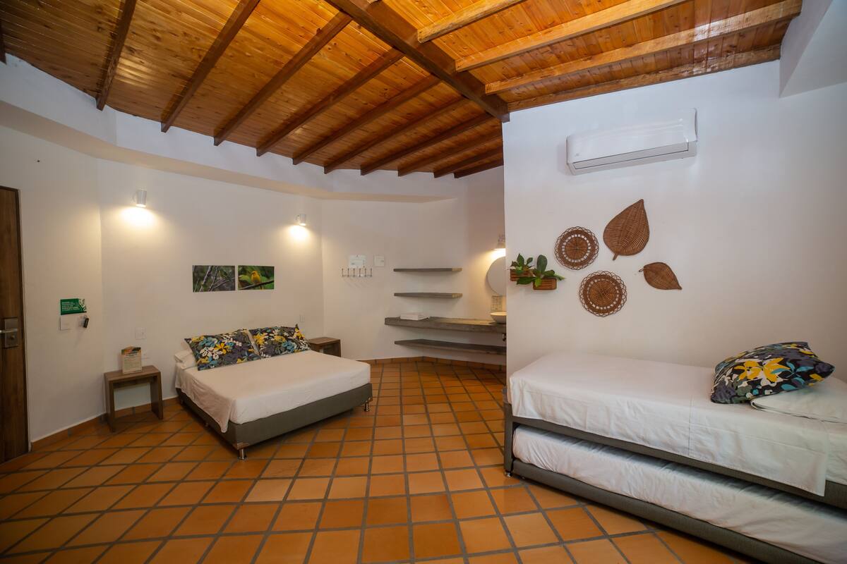 A bedroom with two beds, a wooden ceiling, a tiled floor, a wall-mounted air conditioner, and a small bedside table with a lamp.