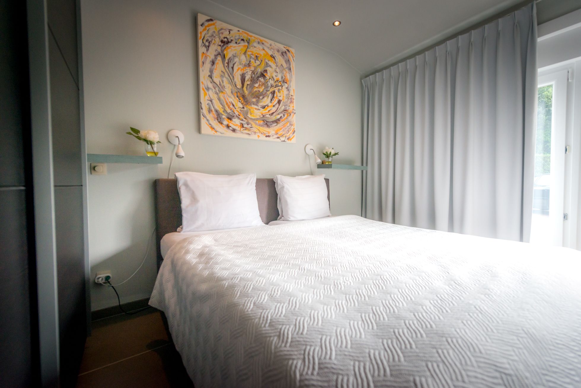 A modern bedroom with a large bed, a decorative abstract painting, and a view of the outdoors through sheer curtains.
