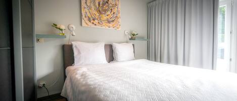A modern bedroom with a large bed, a decorative abstract painting, and a view of the outdoors through sheer curtains.