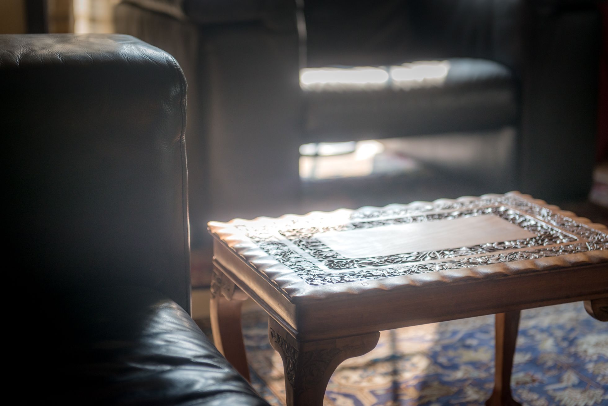 Una pequeña mesa de madera con detalles de metal, ubicada en una habitación con un sillón de cuero y una alfombra estampada.