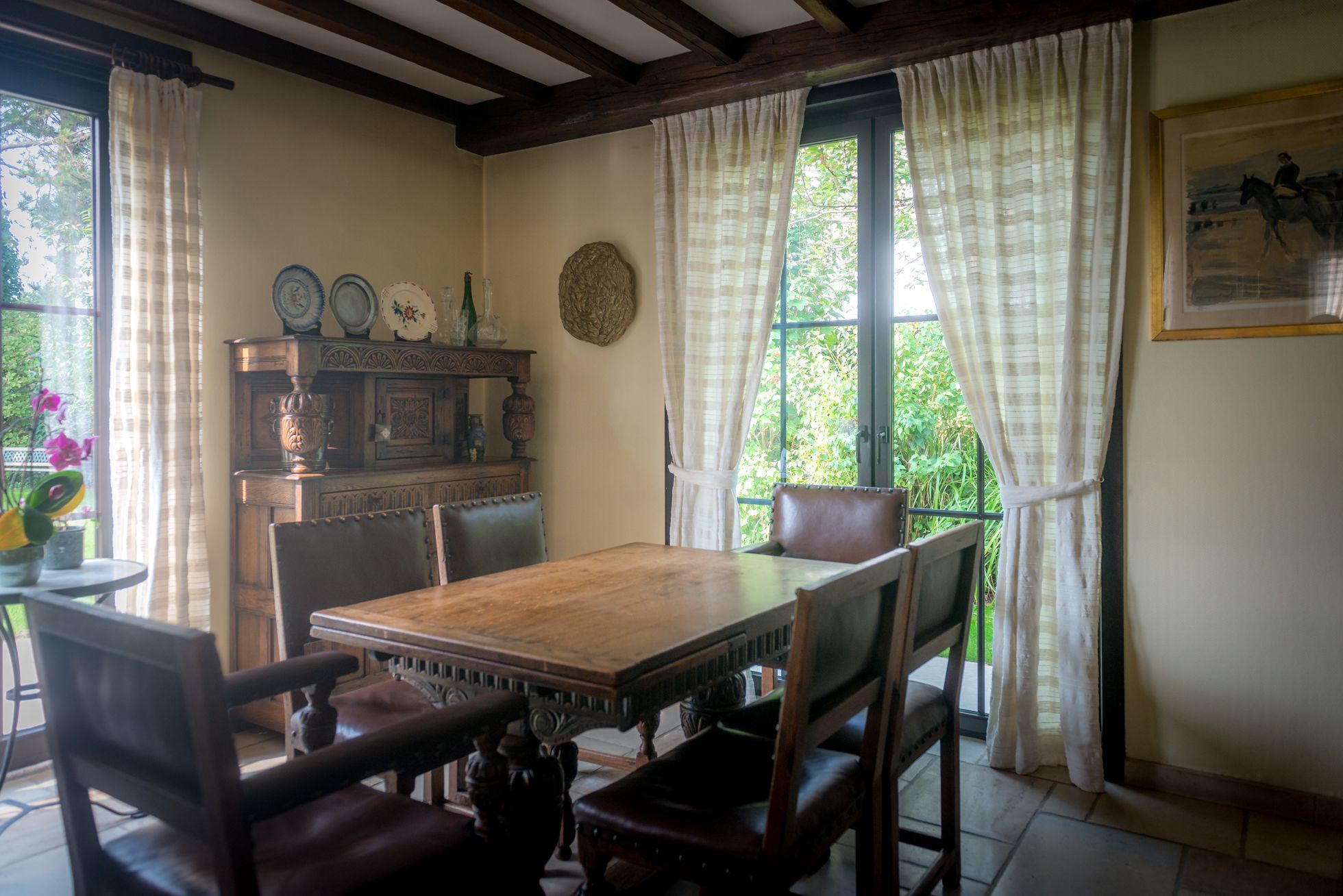 Un coin repas avec une table et des chaises en bois, un buffet orné d’assiettes décoratives et une fenêtre avec des rideaux.