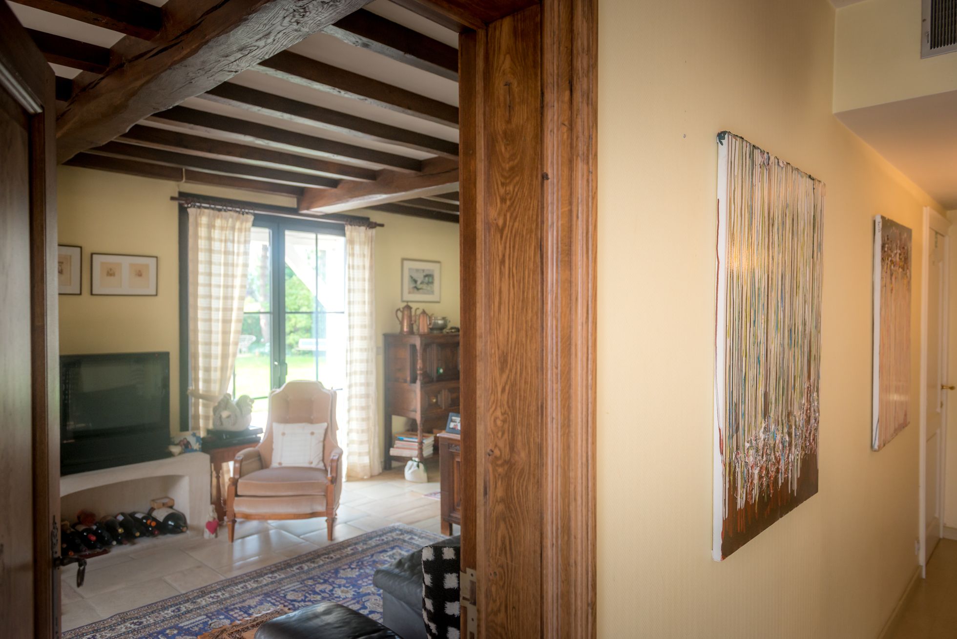 Una habitación con techo de madera, un ventanal con cortinas, una silla, un televisor y un cuadro en la pared.