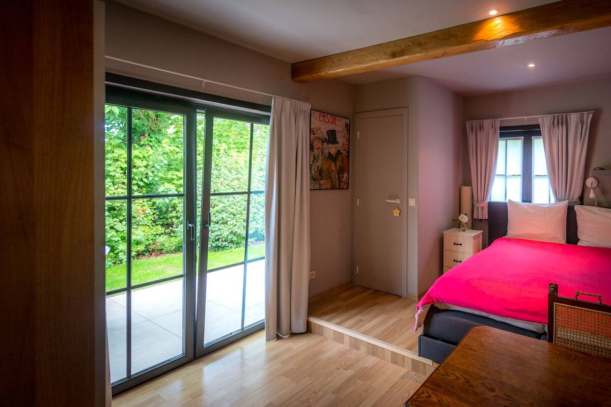 A bedroom with a large window, a bed with a red bedspread, a wooden table, and a bathroom with a shower curtain.