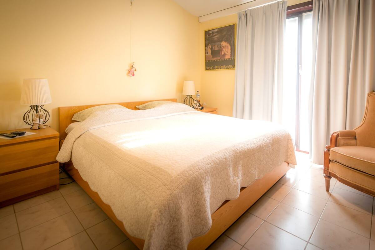 A hotel room with a bed, a nightstand, a chair, and a painting on the wall.