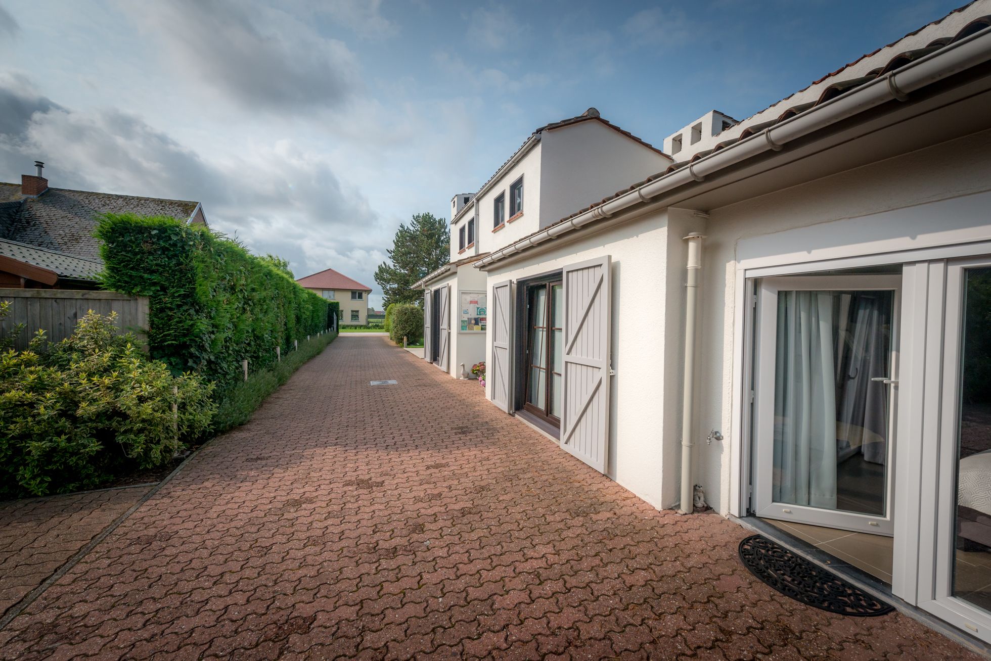 A residential street with a paved walkway, white houses with shutters, and a hedge on the left.