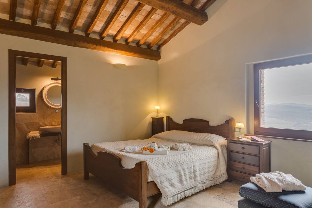 A bedroom with a wooden bed, a nightstand with a lamp, a window with a view, and a bathroom with a round mirror and sink.