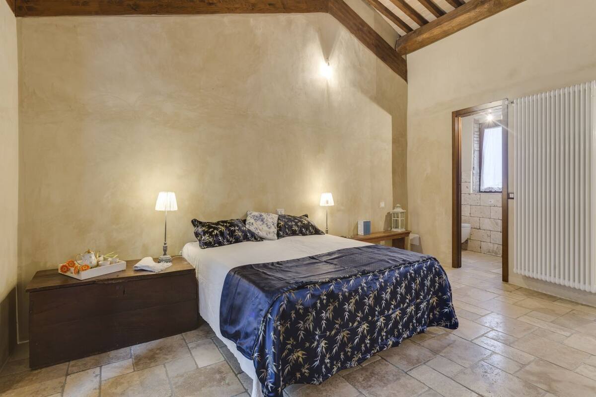 A bedroom with a bed, two bedside tables, a nightstand, a dresser, a window with blinds, and a door leading to another room.