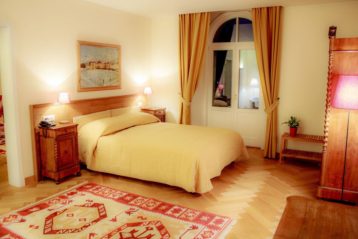 Ein Schlafzimmer mit einem großen Bett, einem Holzkopfende, zwei Nachttischen mit Lampen, einem roten Teppich mit Muster und einem Fenster mit Vorhängen.