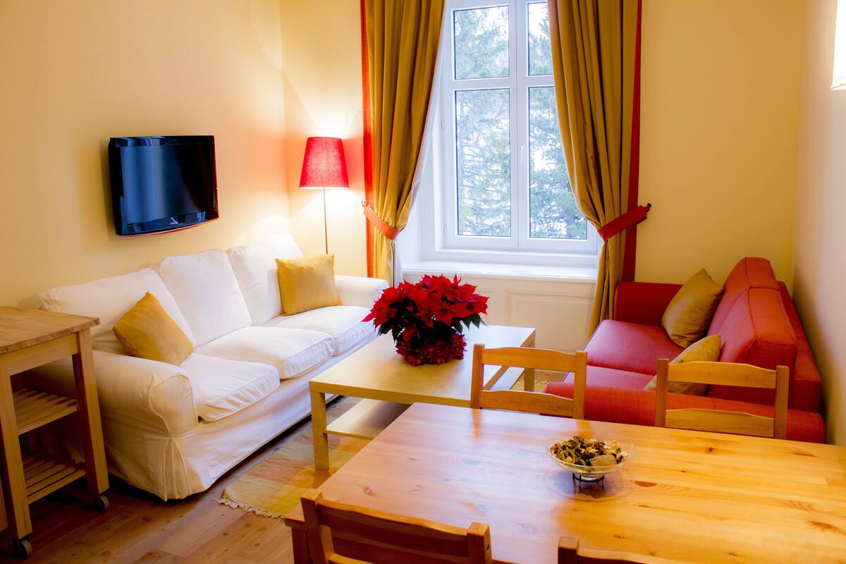 Ein Wohnzimmer mit einem weißen Sofa, einem Holztisch und einem roten Sofa. An der Wand ist ein Fernseher befestigt und es gibt ein Fenster mit Vorhängen.