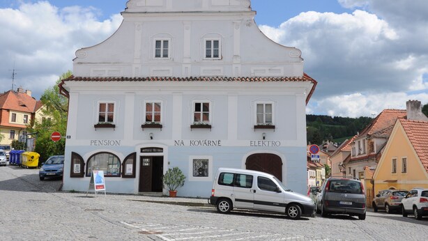 Un edificio blanco con los letreros "PENSION" y "NA KOVARNÉ ELEKTRO", una calle adoquinada y coches estacionados.