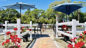 Un espacio al aire libre con bancos y mesas de madera blanca, dos sombrillas y flores rojas.