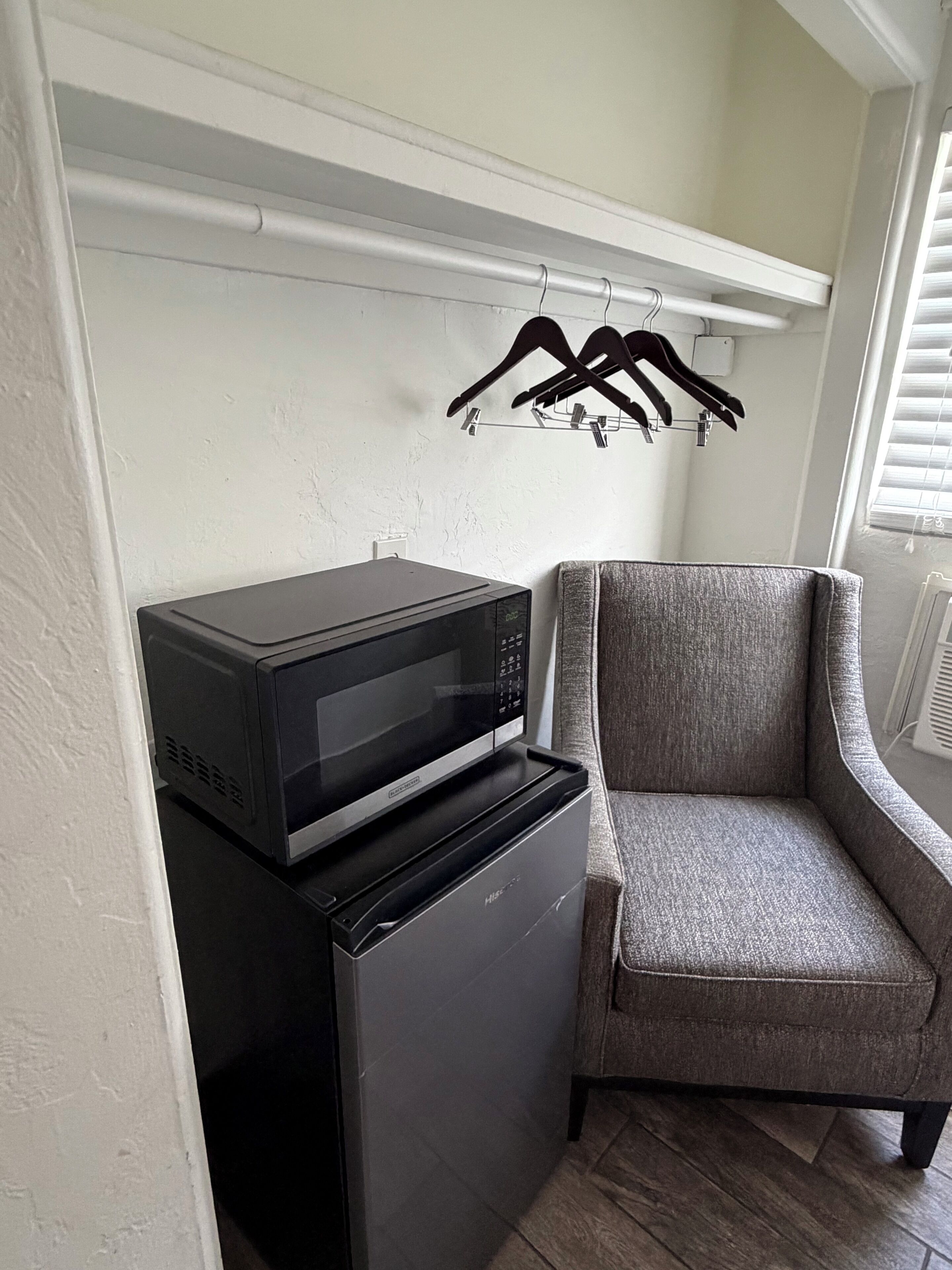 A compact living space with a microwave on a small appliance, a grey upholstered chair, and a clothes rack with hangers.