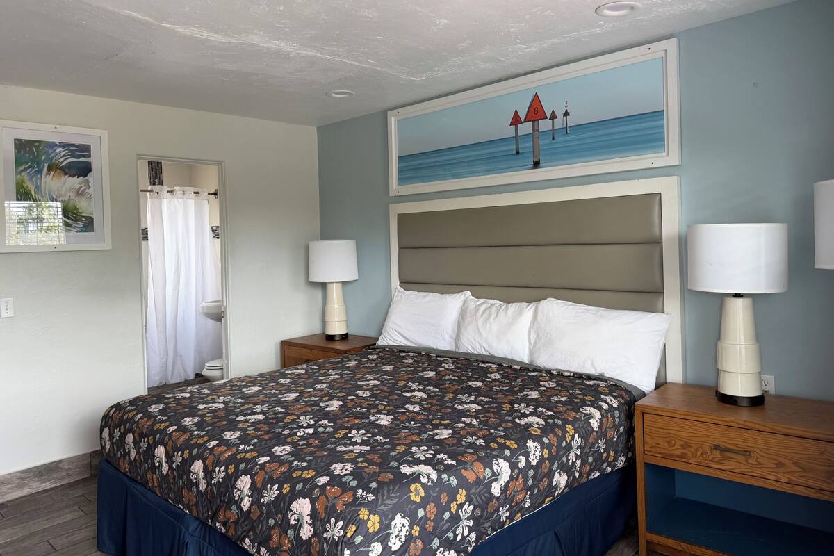 A hotel room with a bed, two bedside tables with lamps, a headboard, a painting of a beach scene, and a window with a curtain.