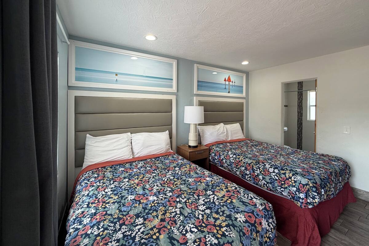 A hotel room with two beds, a nightstand, a lamp, and two framed pictures on the wall.
