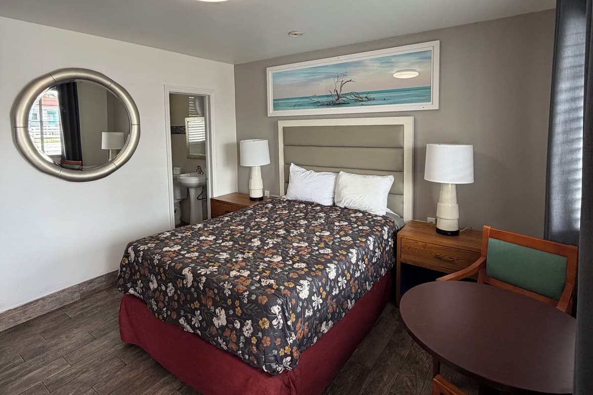 A hotel room with a bed, a round mirror, a painting, a desk with a chair, and a small table.