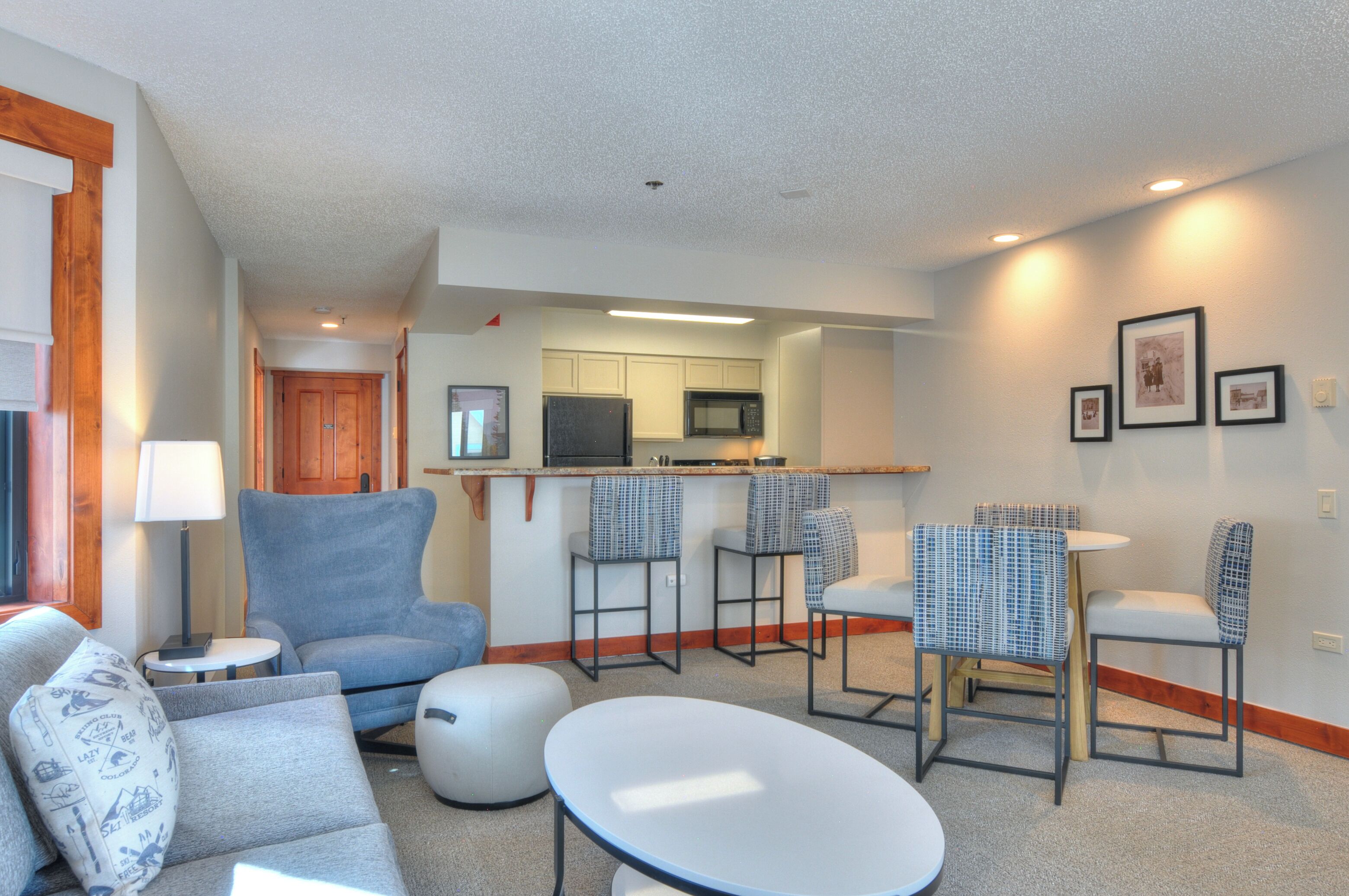 A modern living room with a sofa, ottoman, and a round coffee table. There are two bar stools and a small round table. A kitchen area is visible in the background with a microwave and cabinets.