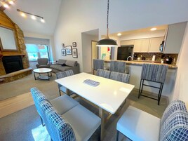 A modern living room with a fireplace, a dining area with a white table and patterned chairs, a kitchen with white cabinets, and a large window providing natural light.