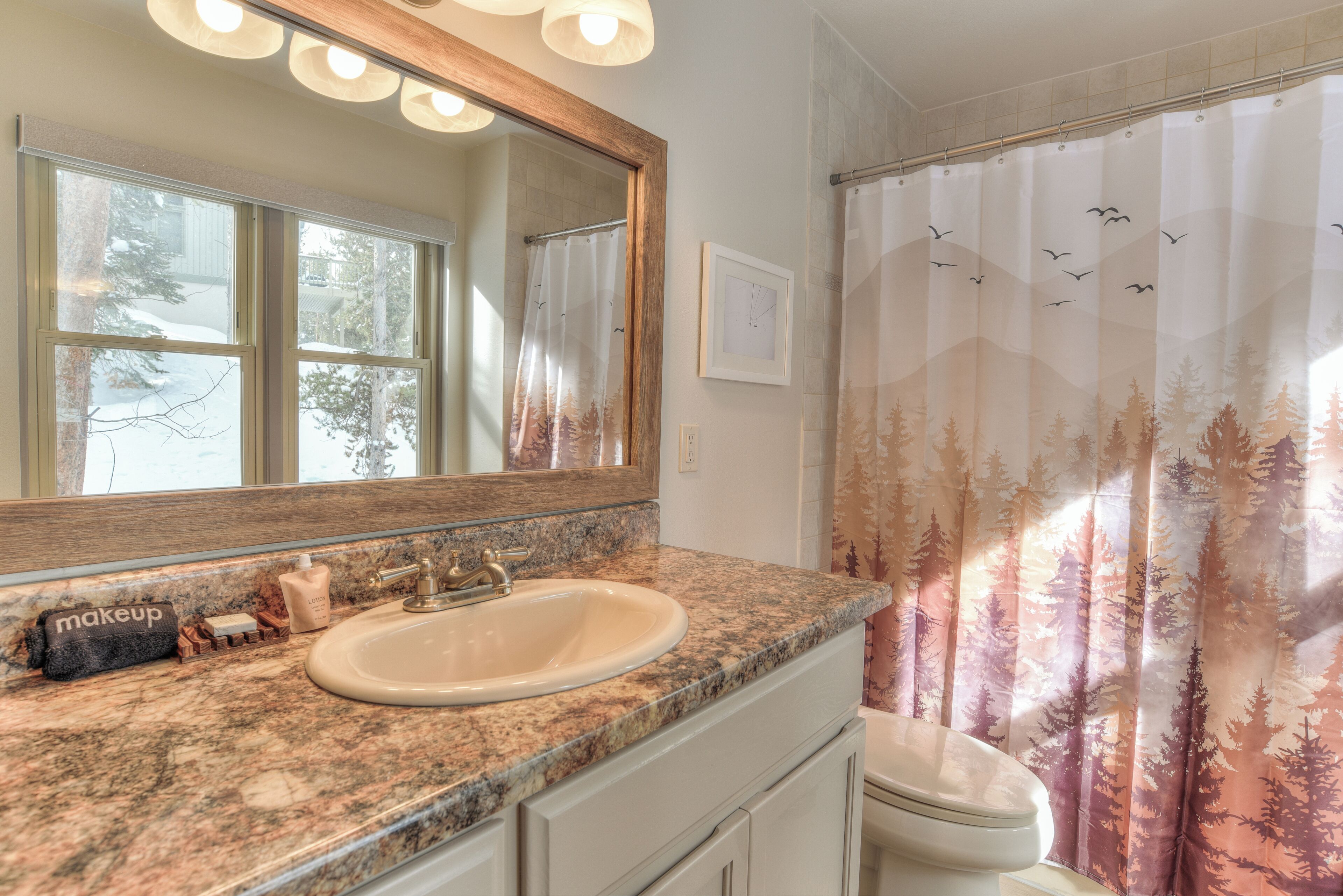 Une salle de bain dotée d’un comptoir en granit, d’un lavabo, d’un miroir et d’un rideau de douche orné d’un motif de forêt.